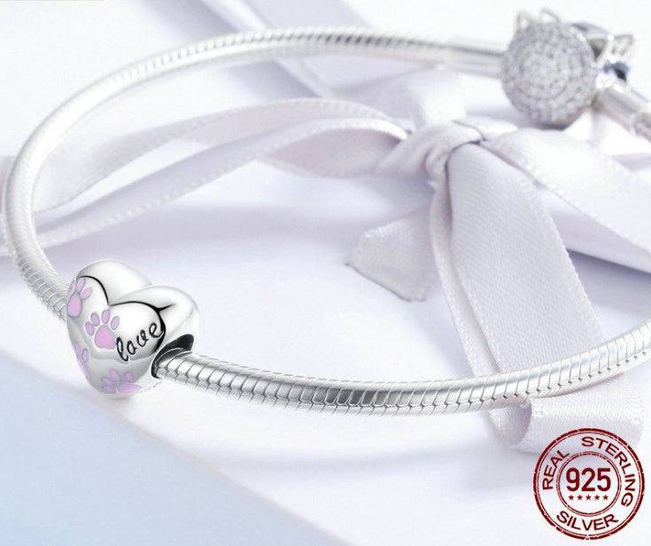 Sterling silver Pink Paw Charm attached to a bracelet, featuring pink paw prints and 'love' engraving. A charming accessory for pet lovers.